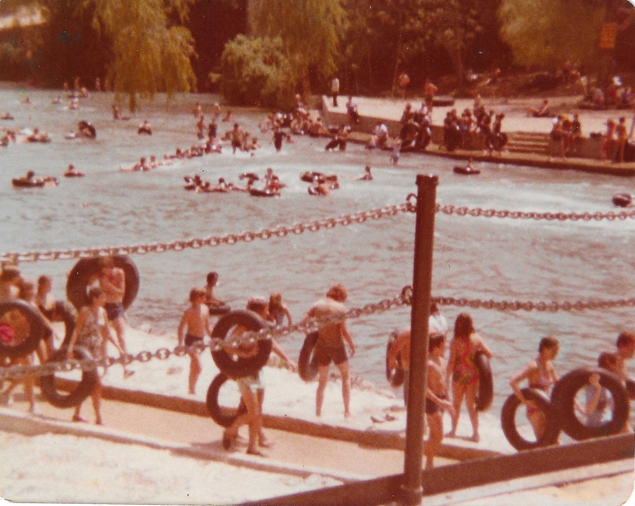 Historical 1977 photograph of tubers walking around the bank of the Comal River Tube Chute with old style black tubes in New Braunfels Texas